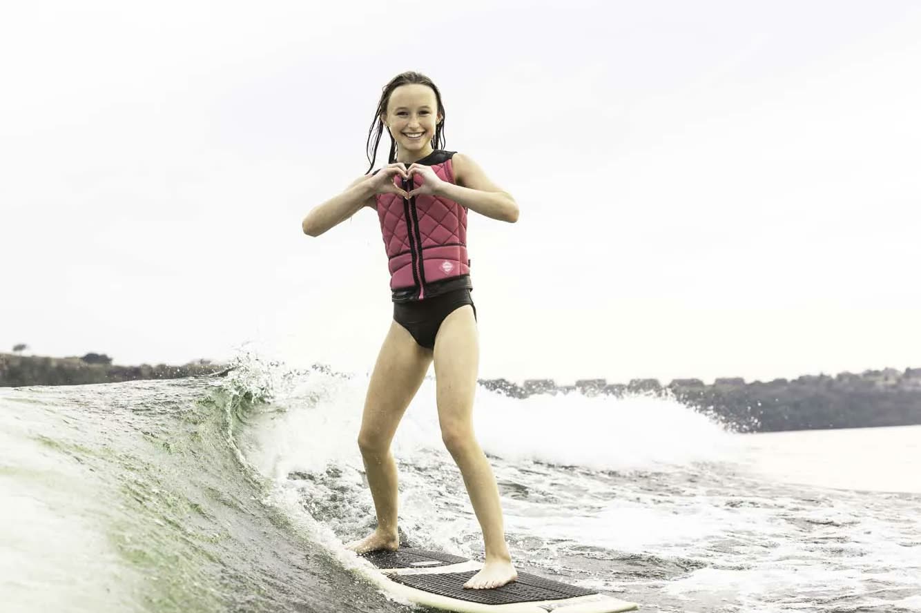 Smiling young rider in a pink life vest making a heart with her hands while wakesurfing behind the boat at Lake of the Ozarks